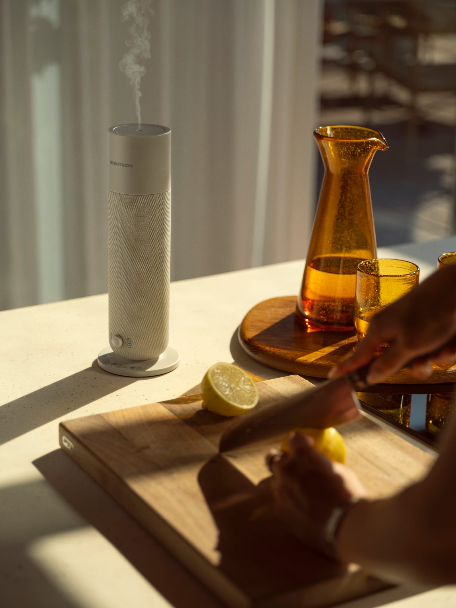 Scentech Smart Aroma Diffuser 2 met nevel op het aanrecht naast een amberkleurige karaf, terwijl iemand een citroen snijdt